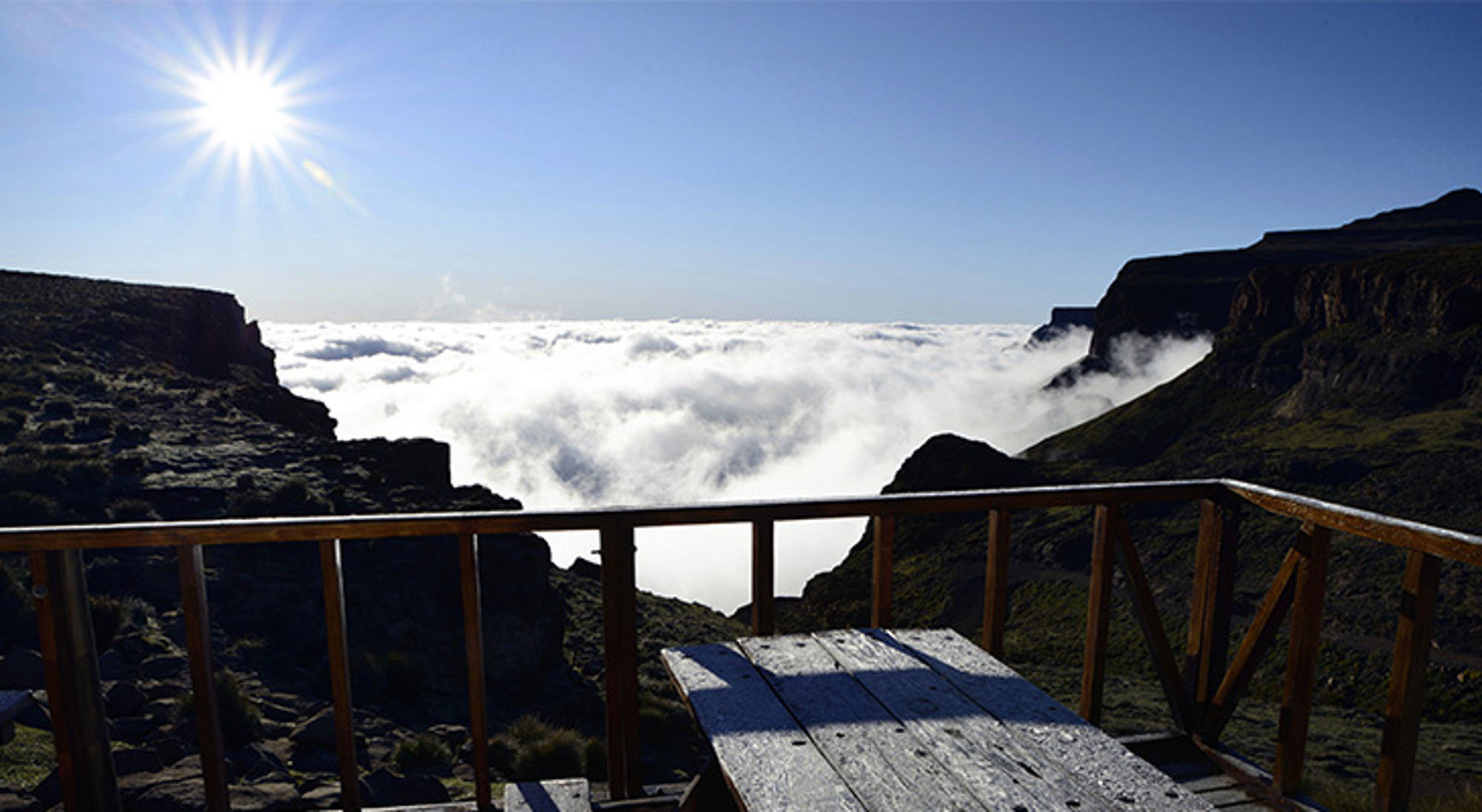 Uitzichtsplek bij Sani Pass Uitzicht vanaf een houten terras over een wolkenzee tussen bergen in Zuid-Afrika, met de zon hoog aan de hemel.