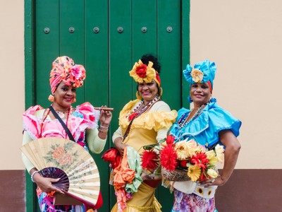 Drie vrouwen in kleurrijke Cubaanse kleding poseren met waaier en bloemenmand voor een groene deur.