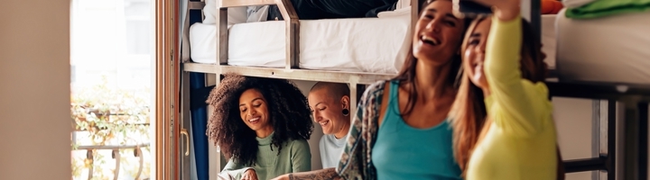 Vrienden in een hostelkamer met stapelbedden; twee maken een selfie terwijl anderen ontspannen.