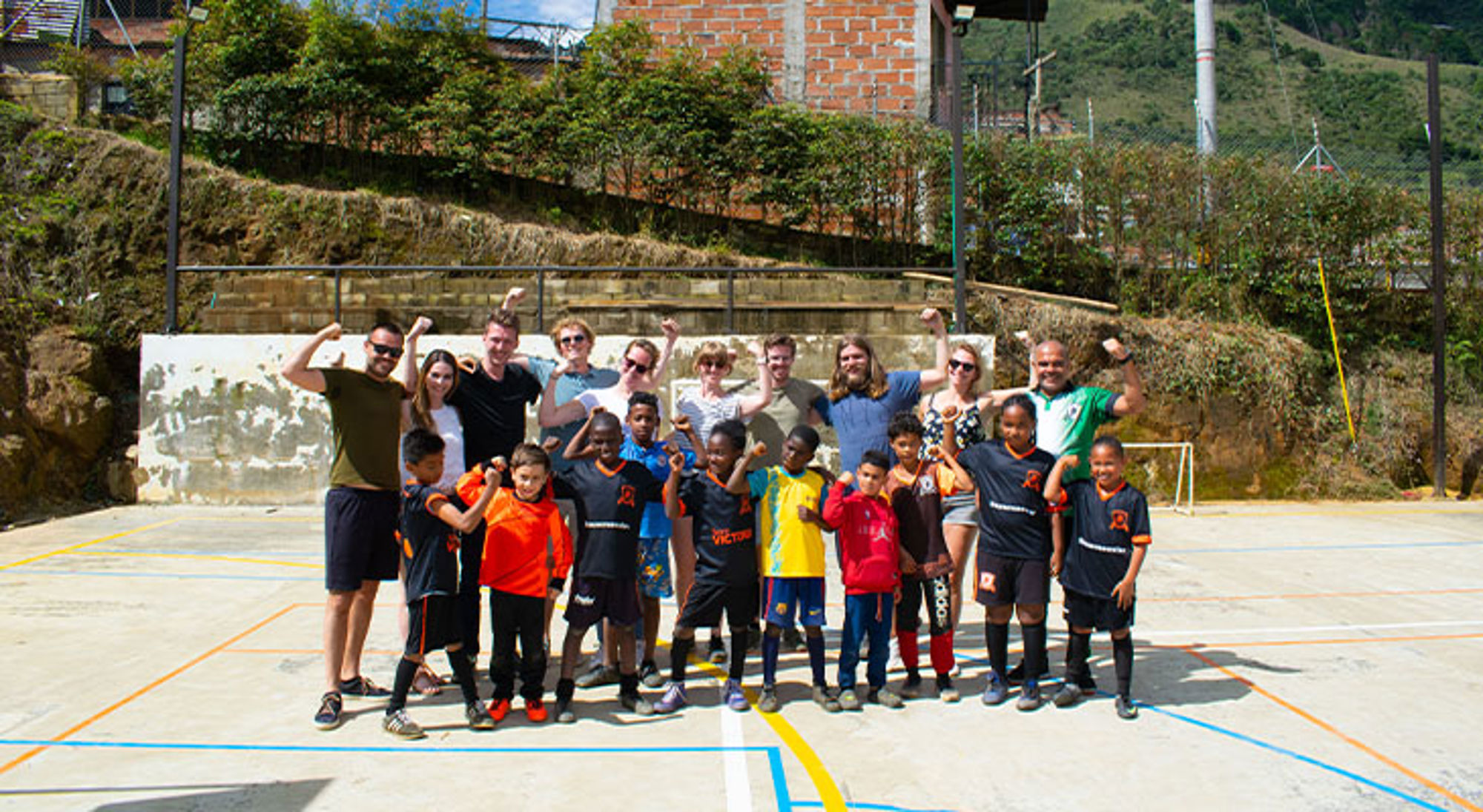 Sawadee reizigers bezoeken Stichting Children of Medellín Groepsfoto van kinderen in voetbaltenue en begeleiders op een sportveld, met heuvels en huizen op de achtergrond.