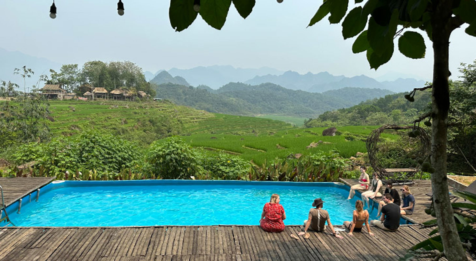 Zwembad homestay Vietnam Zwembad met reizigers op houten terras met uitzicht op groene rijstvelden en bergen.