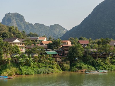 Rivierdorp aan de Mekong in Laos, met groene bergen op de achtergrond en boten op het water.