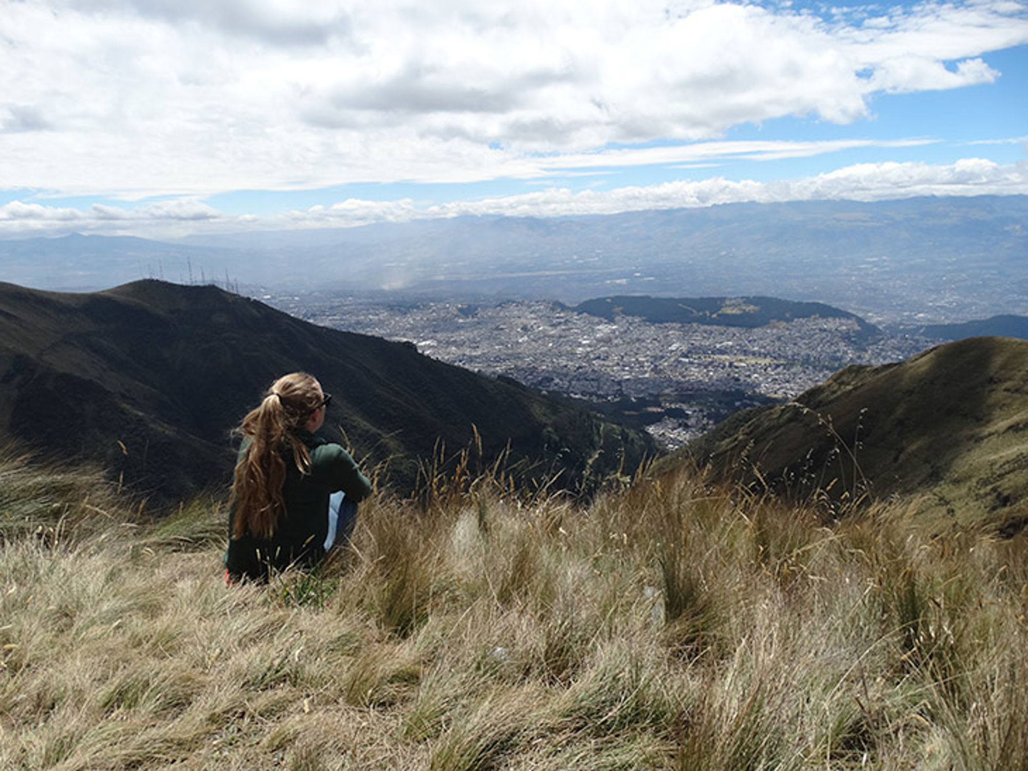 Pichincha berg, Ecuador Wandelaar kijkt uit over een vallei en stad tussen groene bergen onder een bewolkte lucht.