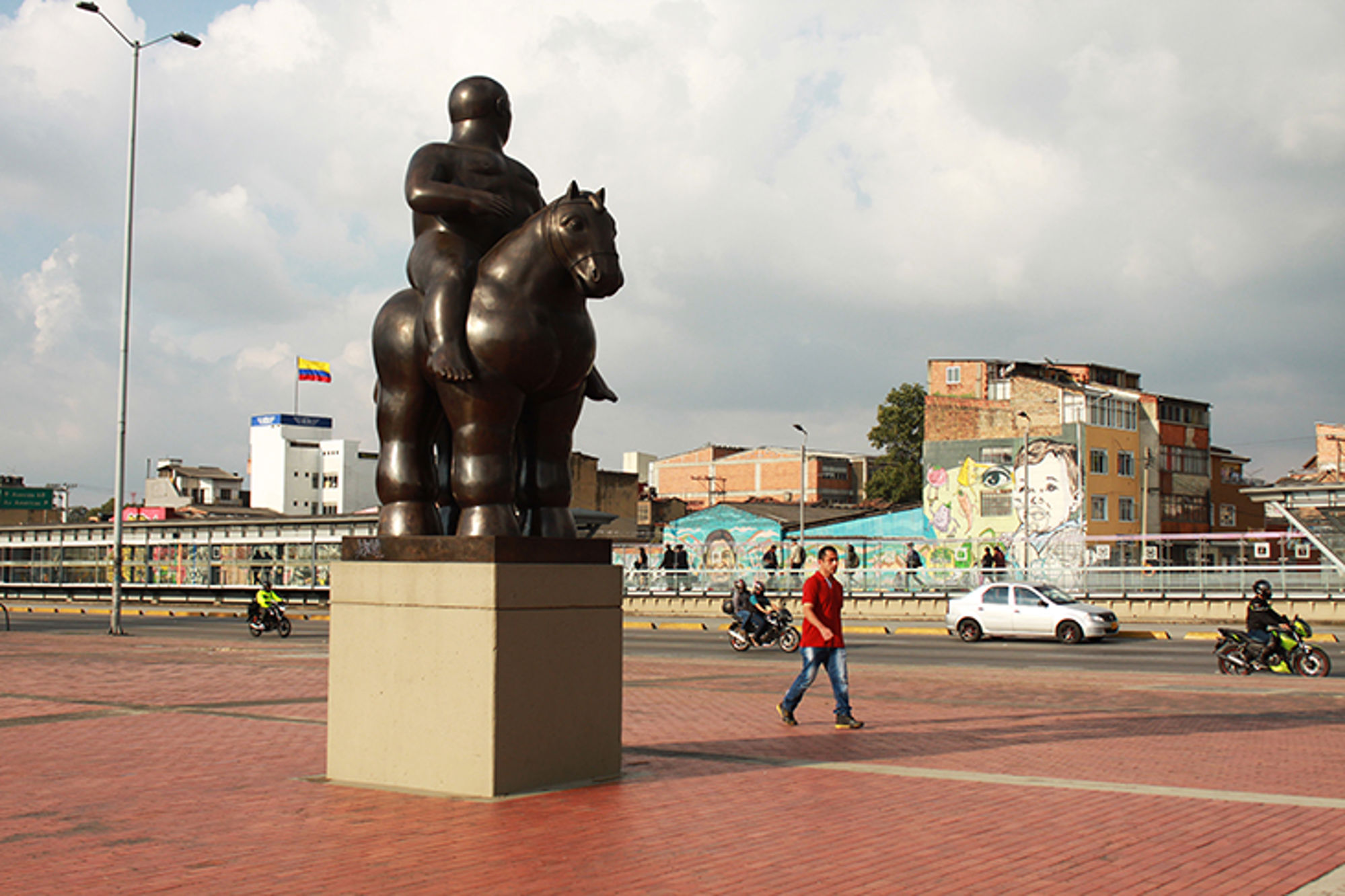 Bronzen standbeeld van een ruiter op een paard op een plein in Medellín, met straatkunst en verkeer op de achtergrond.