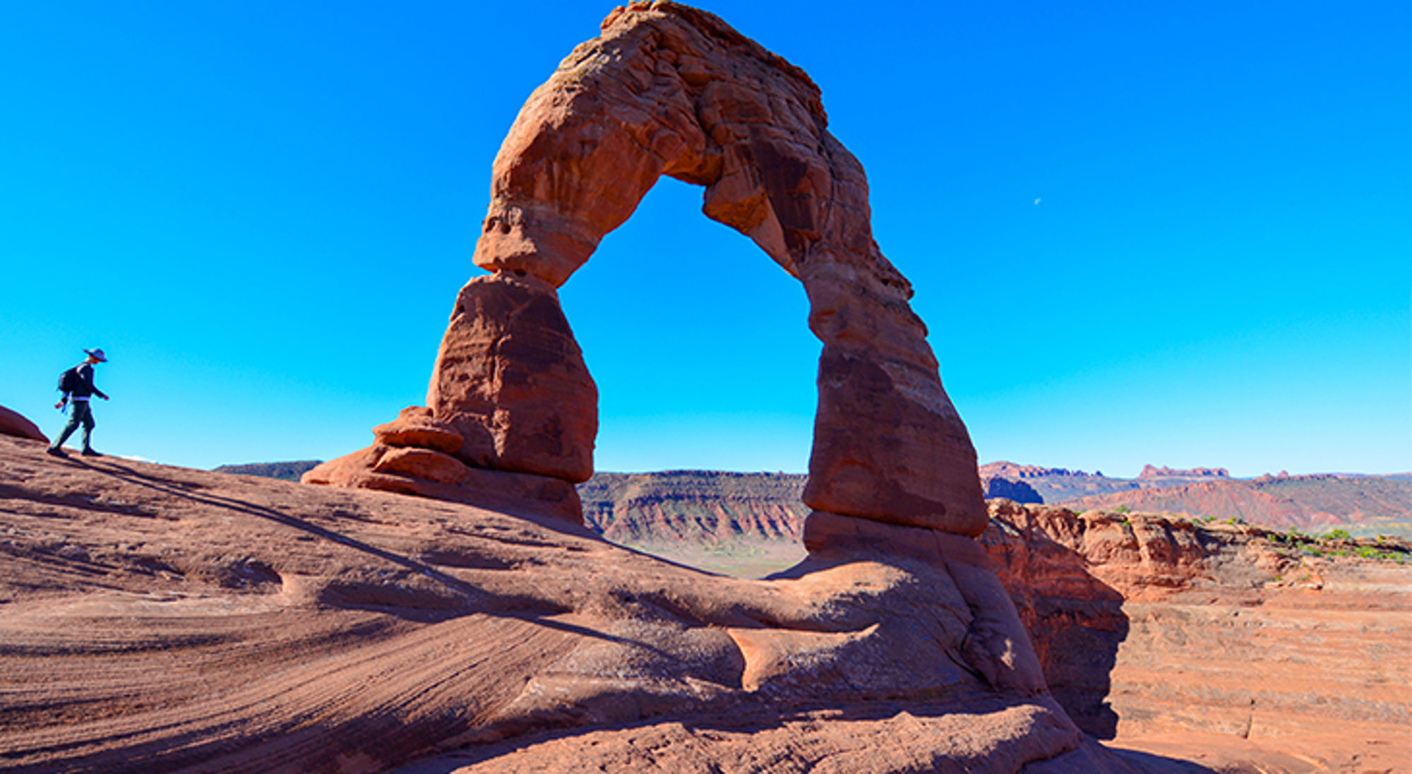 Wandelaar bij Delicate Arch in Arches National Park, Utah, tegen een heldere blauwe lucht.