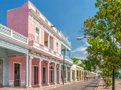 Pastelkleurige koloniale straat in Cuba met arcades, balkons en groene bomen langs de weg.