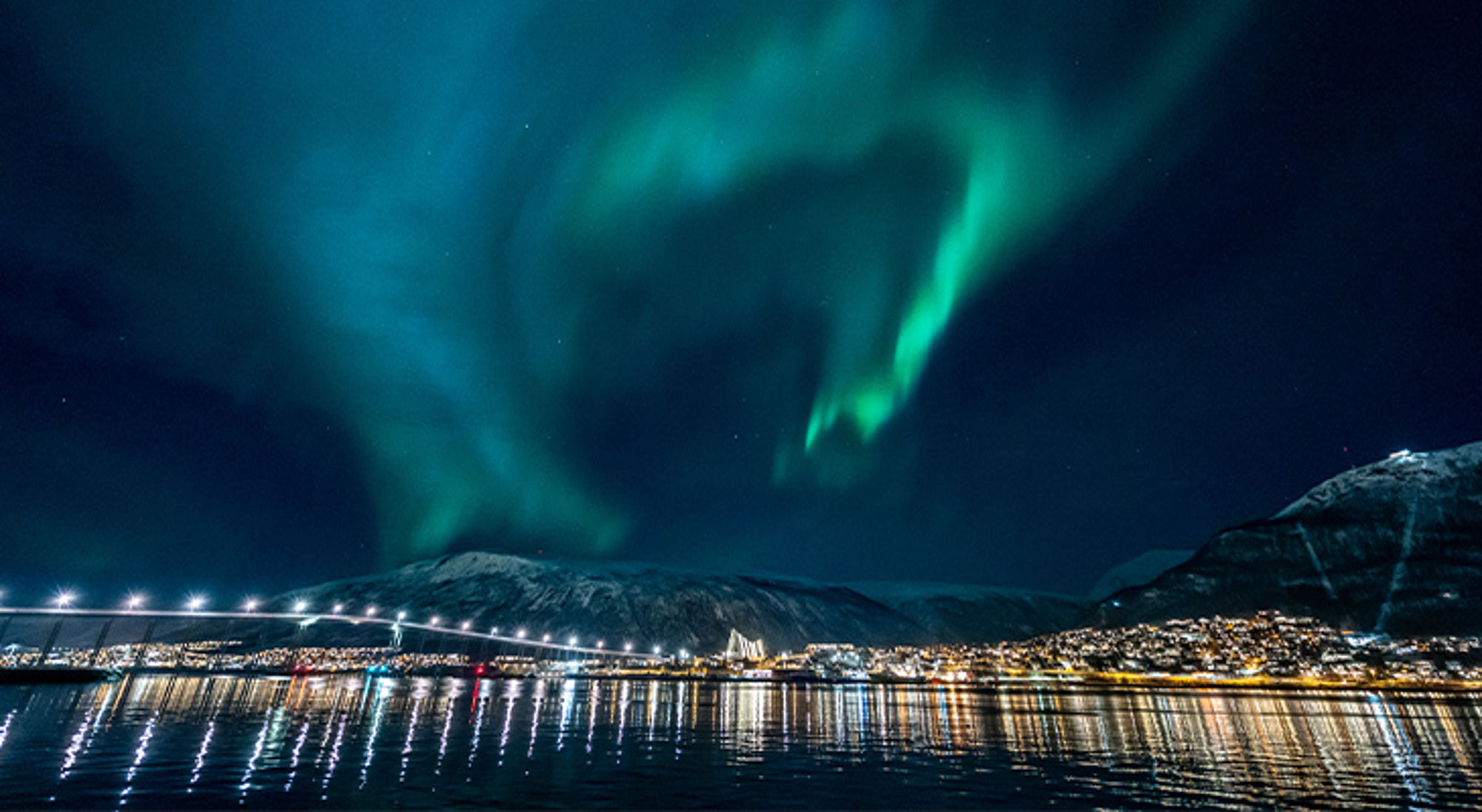 Noorderlicht in Tromsø, Noorwegen Noorderlicht boven een verlichte kuststad met brug en weerspiegeling in het water.