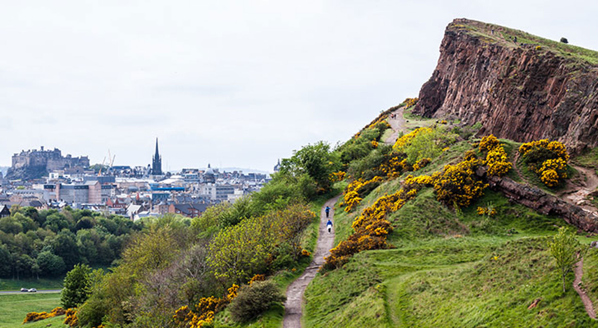 De mooiste stad ter wereld Edinburgh Uitzicht vanaf Arthur’s Seat op Edinburgh met wandelpad langs groene heuvels en gele brem.