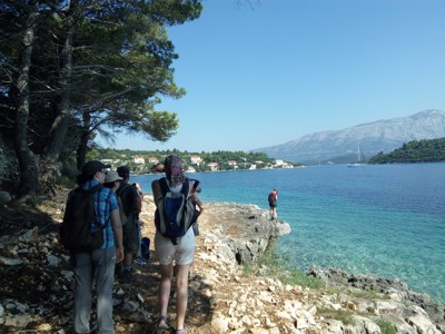 Wandelaars met rugzak langs de Adriatische kust in Kroatië, met helderblauw water en bergen op de achtergrond.