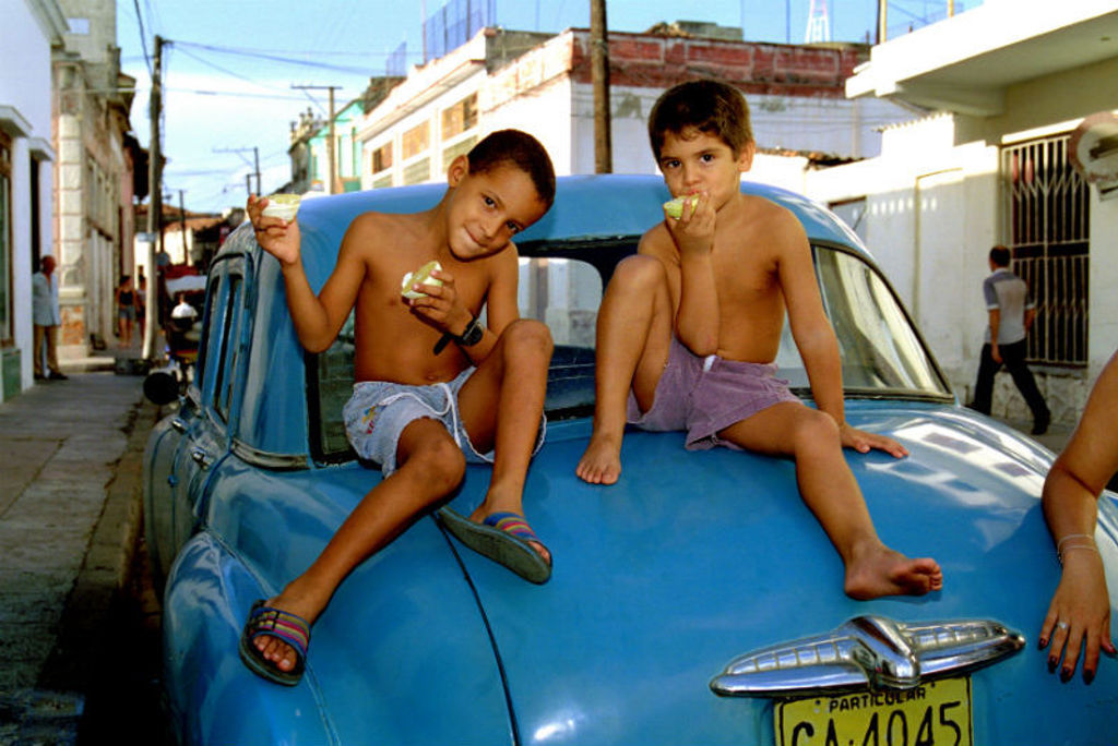Jongens eten fruit op de motorkap van een blauwe oldtimer in een straat in Cuba.
