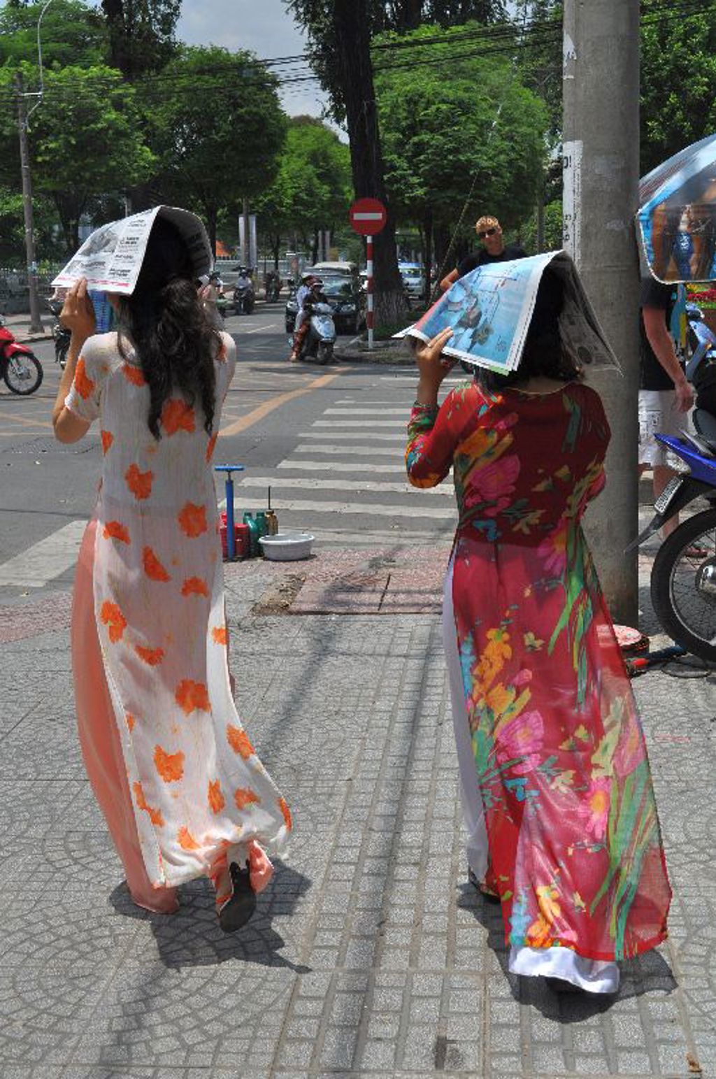 Vrouwen in ao dai steken een straat over in Vietnam en beschermen zich tegen de zon met kranten.