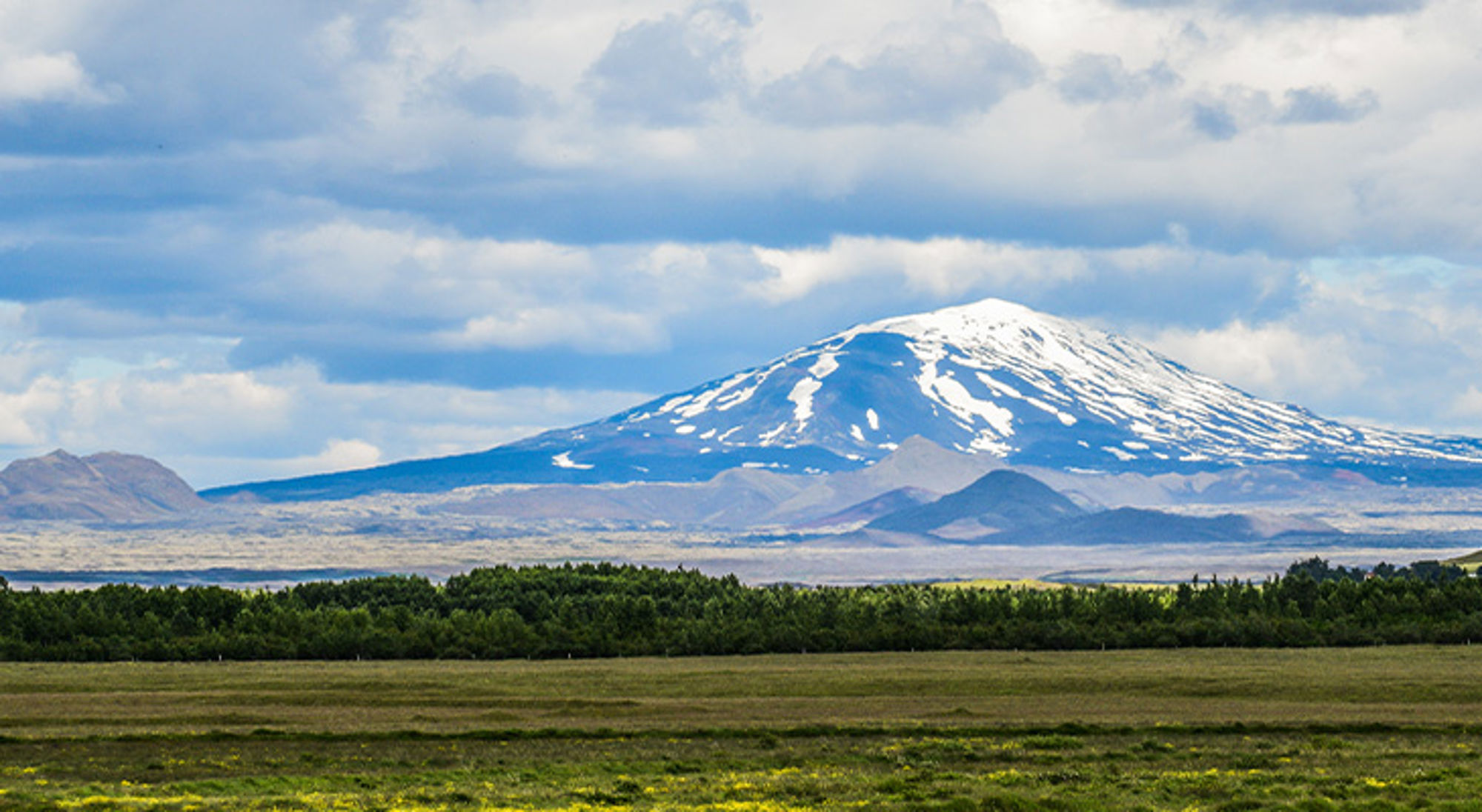 Hekla vulkaan IJsland Sneeuwbedekte bergtop boven een groene vlakte onder een bewolkte lucht.
