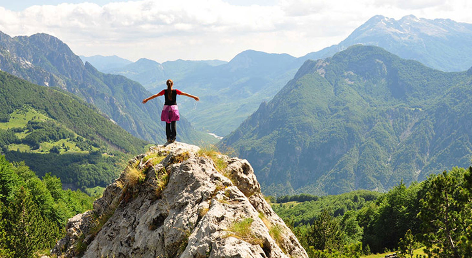 Wandelvakantie Albanië Wandelaar staat met gespreide armen op een rots met panoramisch uitzicht over groene bergen en valleien.