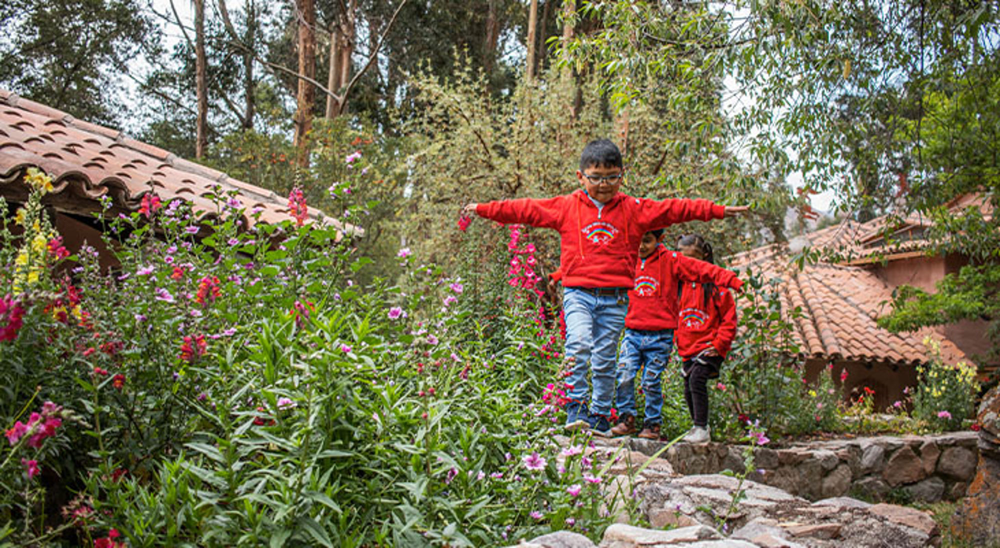 Niños del Arco Iris Kinderen in rode truien balanceren op stenen pad tussen bloemen en huisjes met terracottadaken.