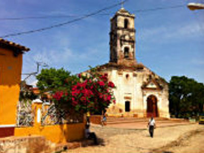 Kerk met klokkentoren aan een dorpsplein op Cuba, met kleurrijke huizen en bloeiende struiken.