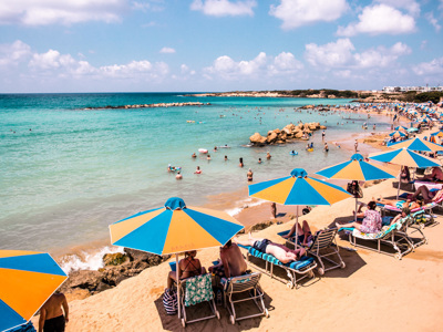 Druk strand op Cyprus met turquoise zee, rotsen en blauw-gele parasols langs de kust.