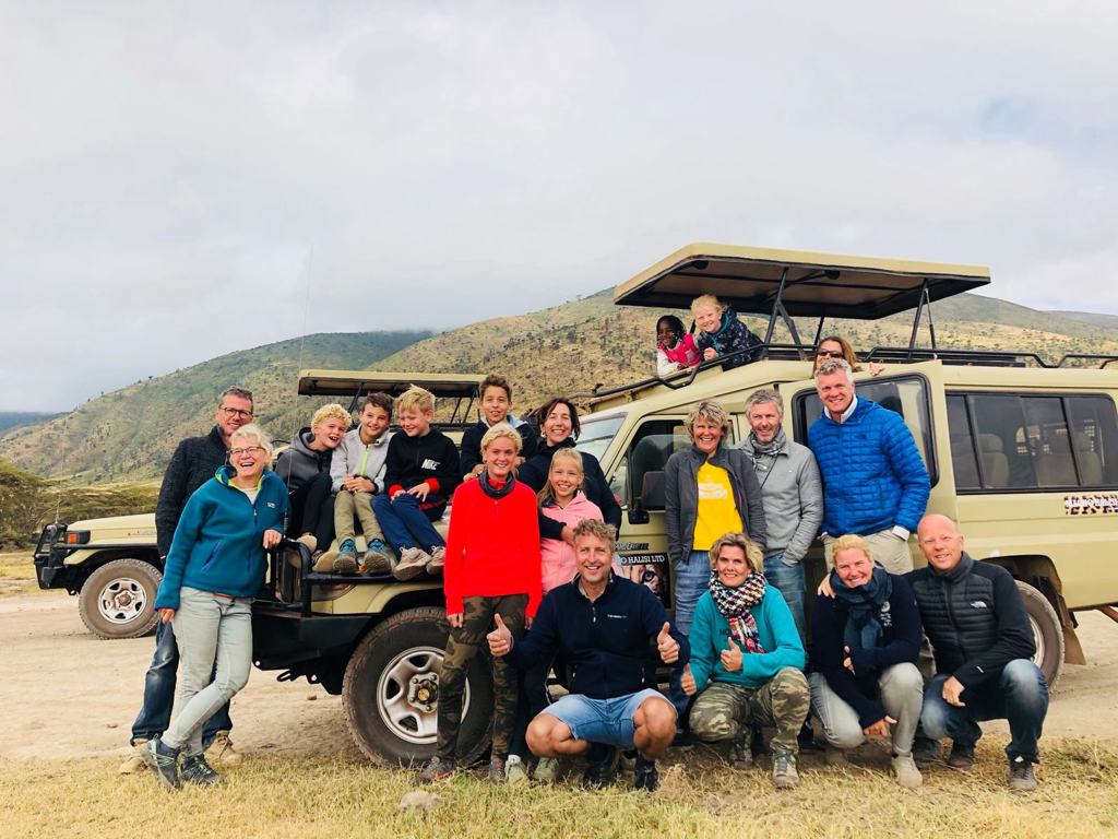 Familiegroep bij safari-jeep tijdens familiereis Tanzania en Zanzibar, met heuvelachtig savannelandschap op de achtergrond.