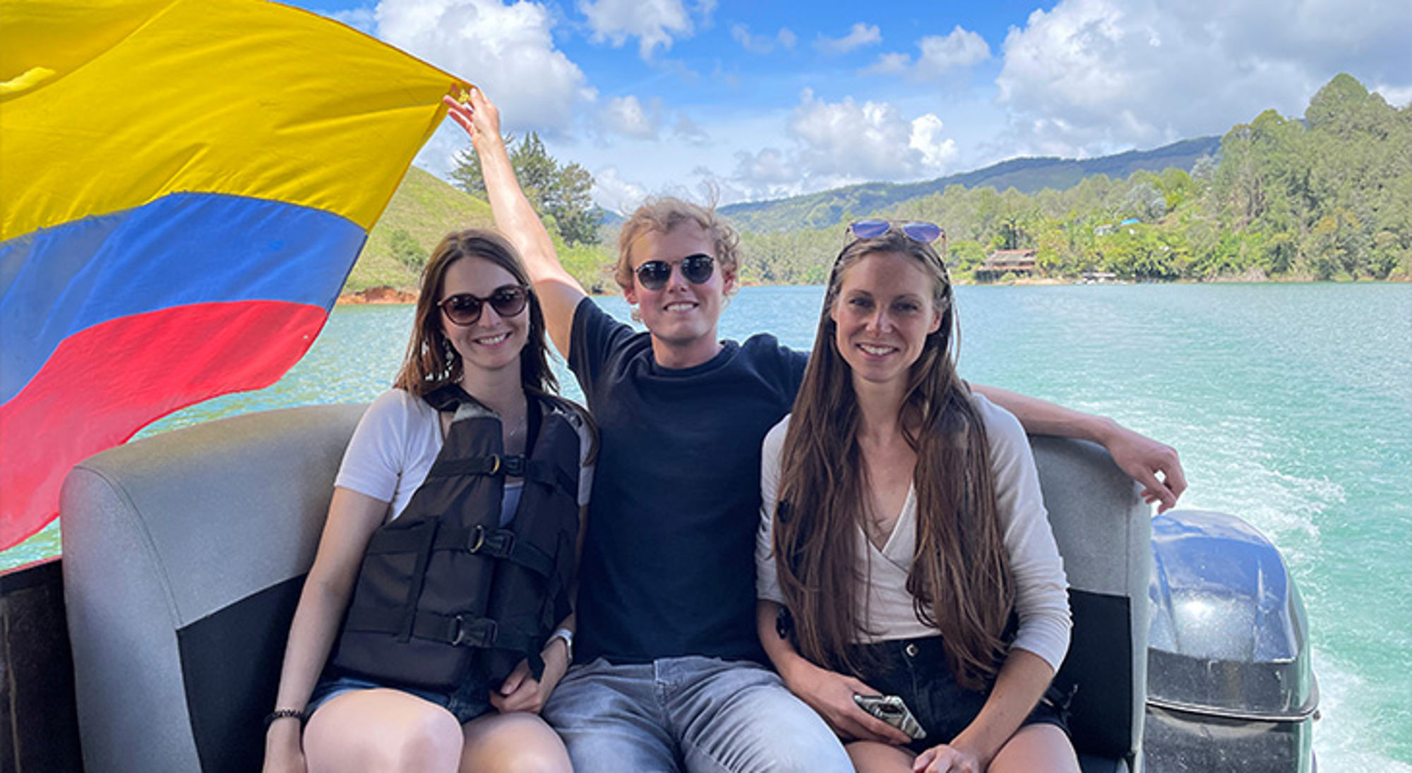 Drie reizigers op een boot op een turquoise meer, met de Colombiaanse vlag en groene bergen op de achtergrond.