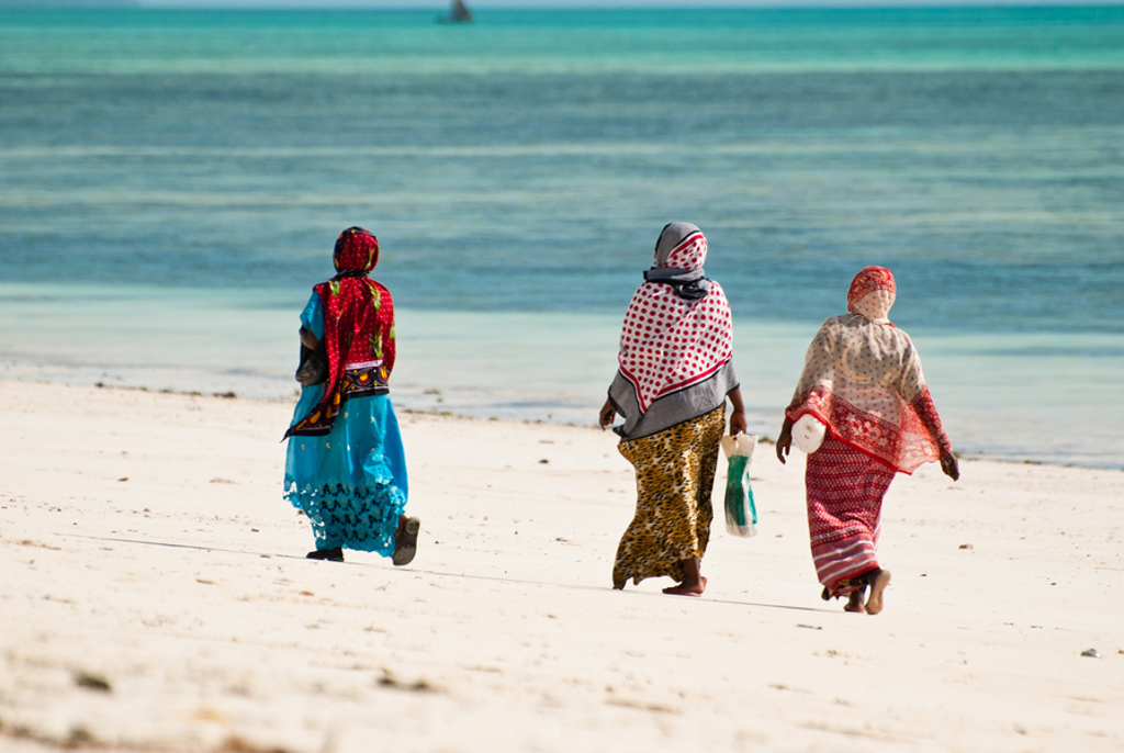 Drie vrouwen wandelen over het witte zandstrand van Zanzibar langs de turquoise zee.