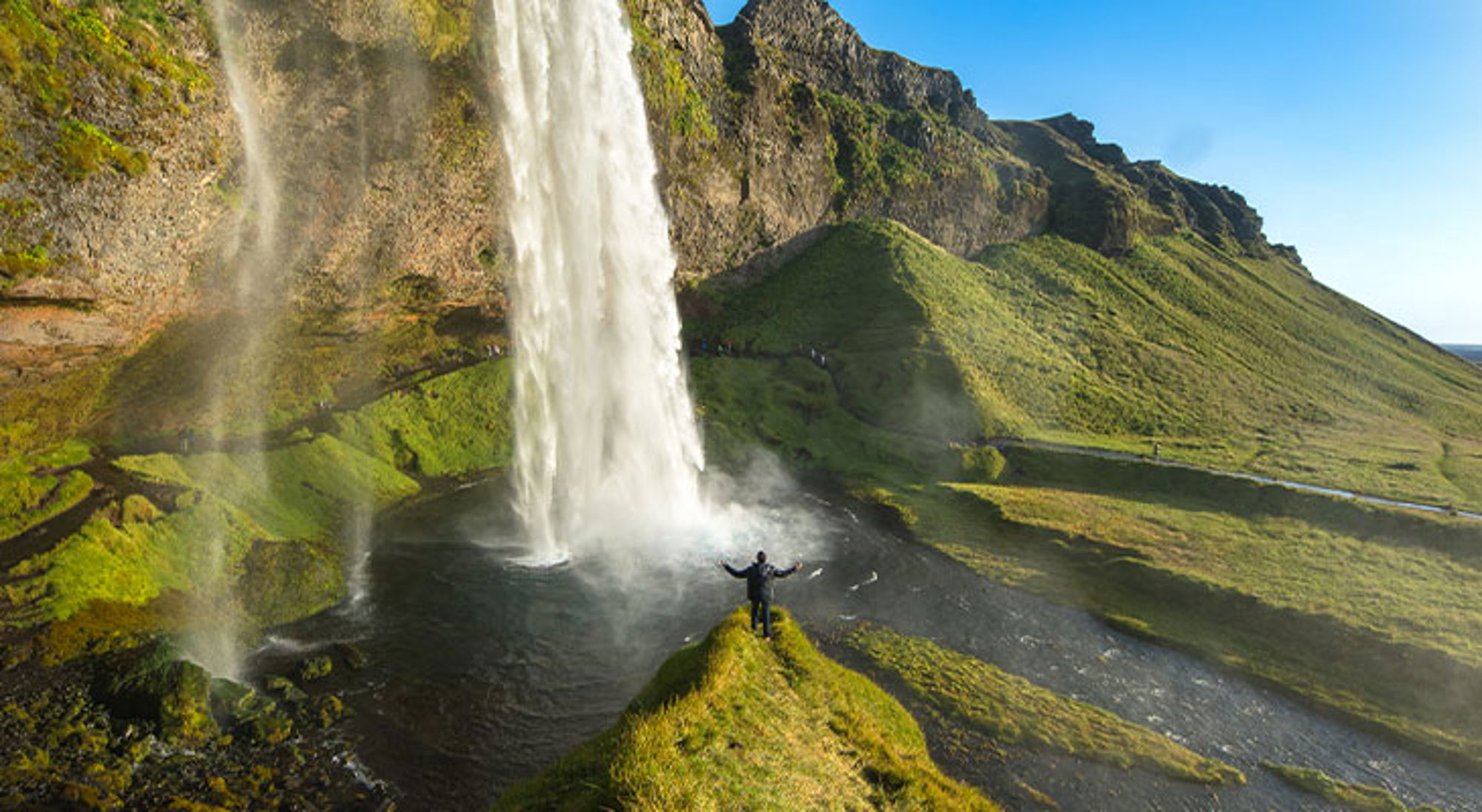 Seljalandsfoss waterval Persoon met gespreide armen bij een hoge waterval in een groen berglandschap.