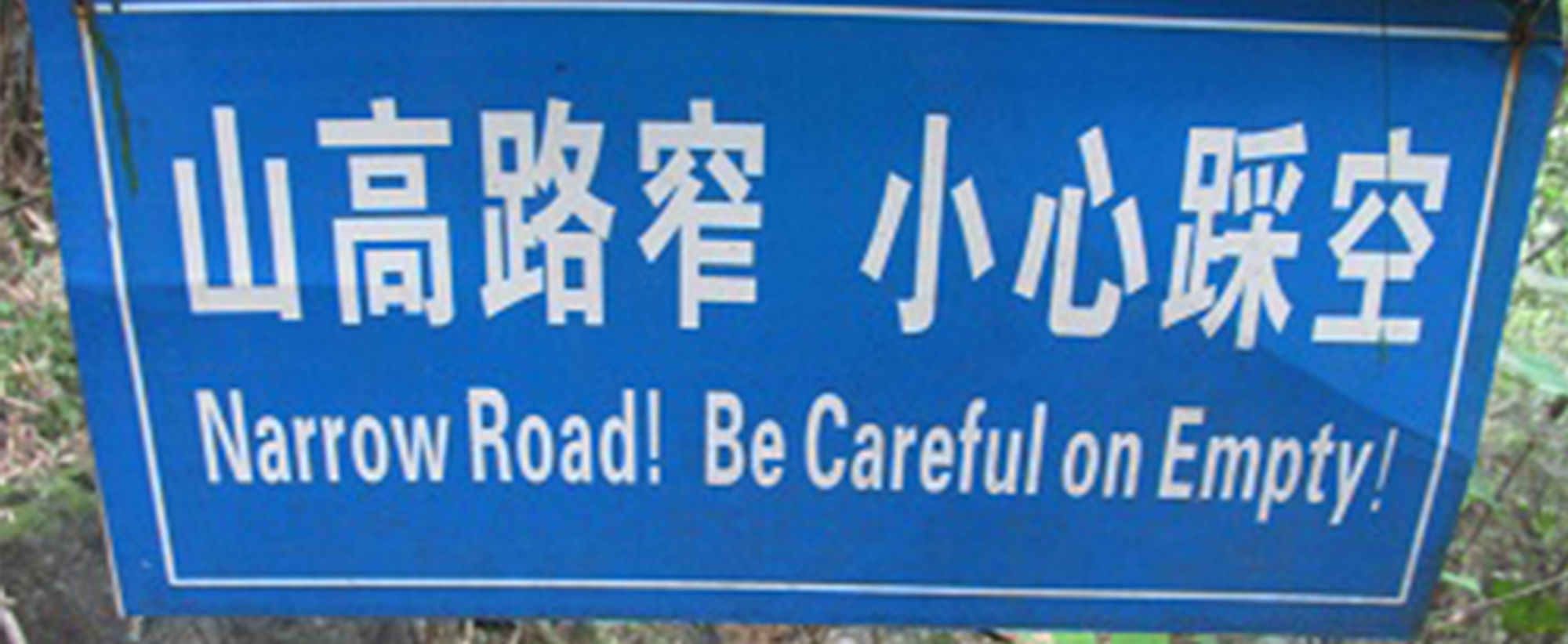 Blauw waarschuwingsbord met Chinese tekens en tekst: “Narrow Road! Be Careful on Empty!”.