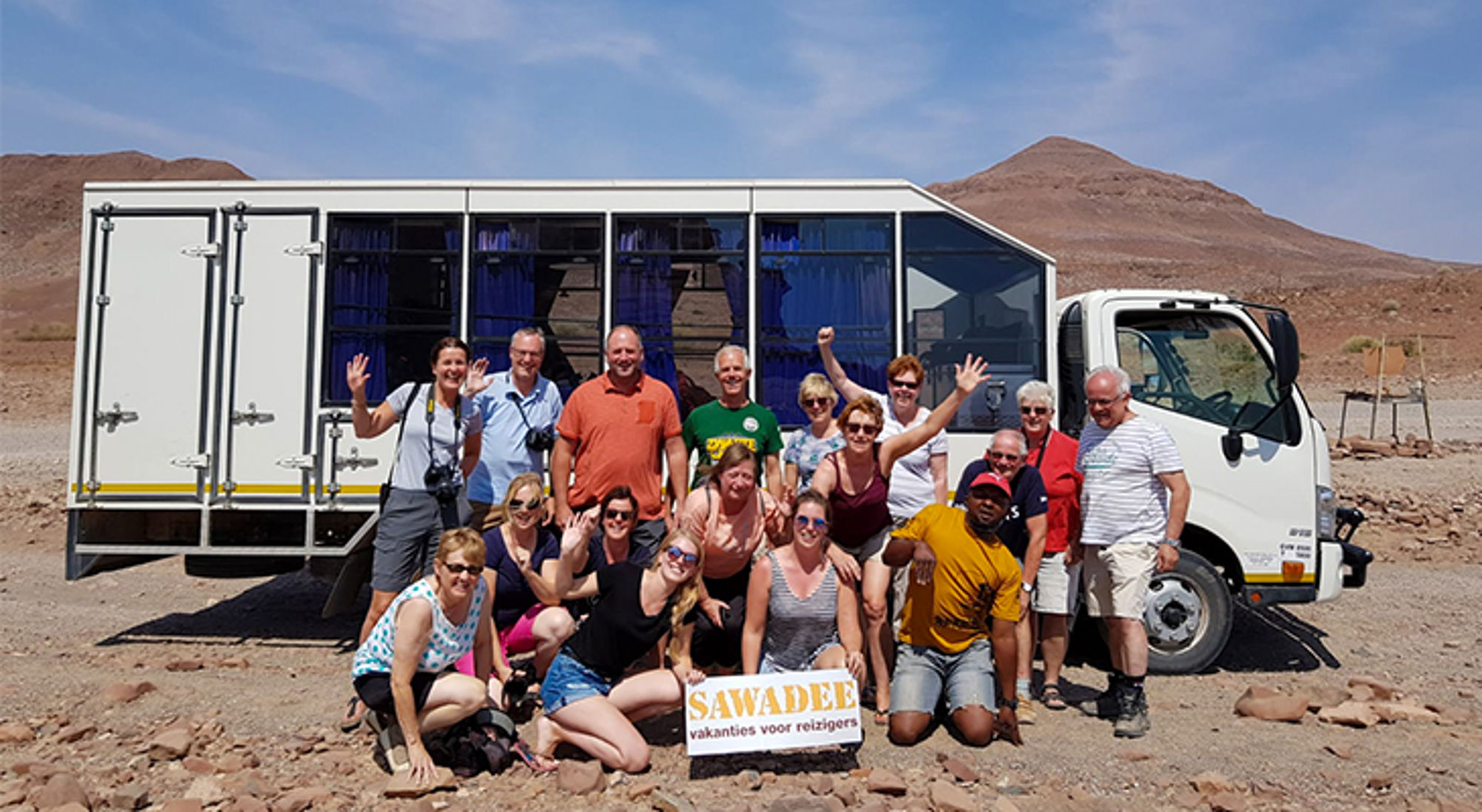 Groep in Namibië Groepsfoto van reizigers bij een truck in een woestijnlandschap, met SAWADEE-bord op de voorgrond.