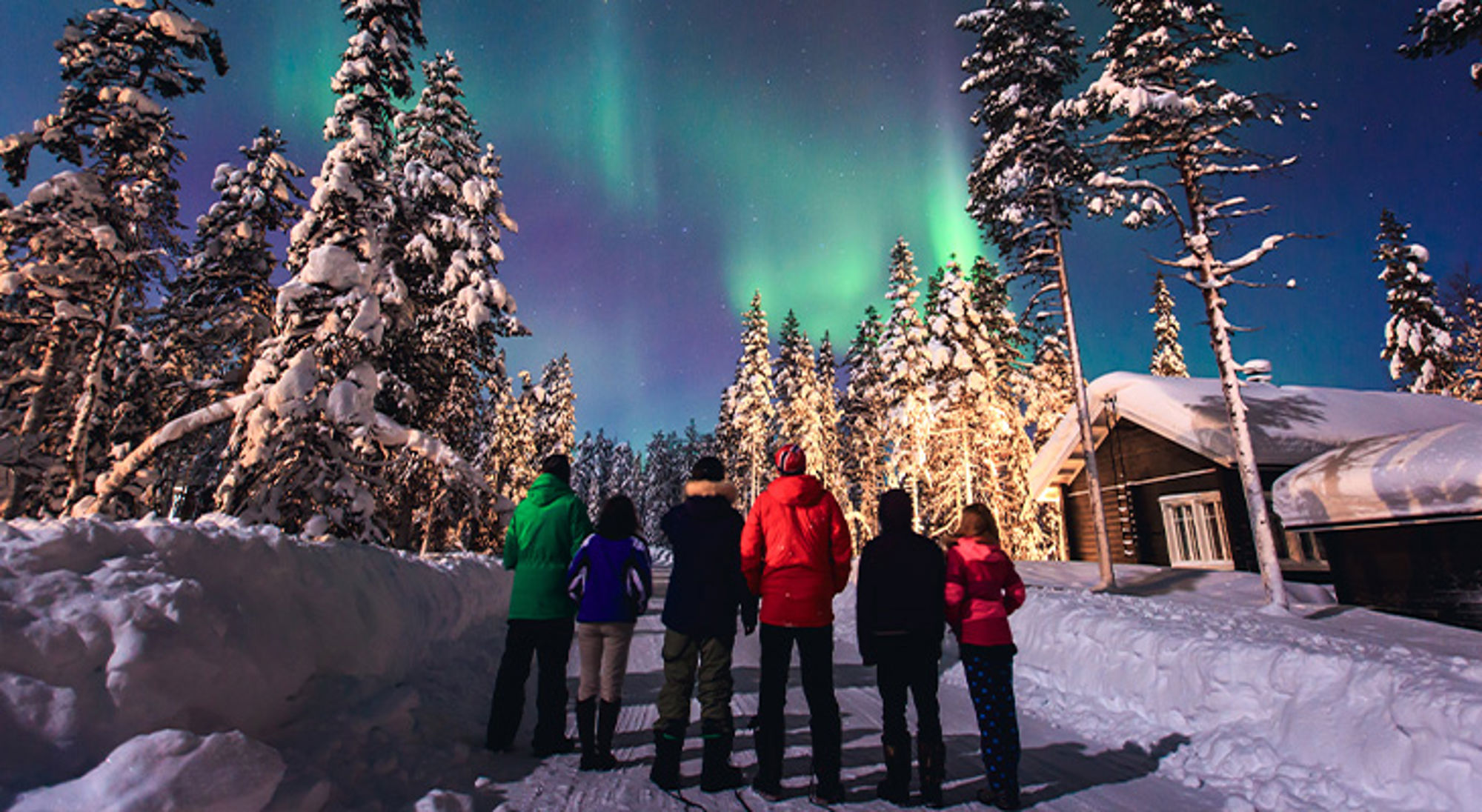 Noorderlicht Lapland Groep reizigers kijkt naar het noorderlicht boven een besneeuwd bos en een houten lodge.