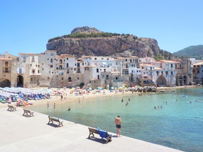 Strand en haven van Cefalù op Sicilië, met historische huizen aan zee en rots van La Rocca op de achtergrond.
