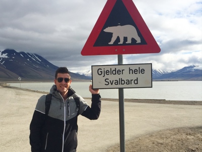 Reiziger bij waarschuwingsbord voor ijsberen op Spitsbergen (Svalbard), met fjord en bergen op de achtergrond.
