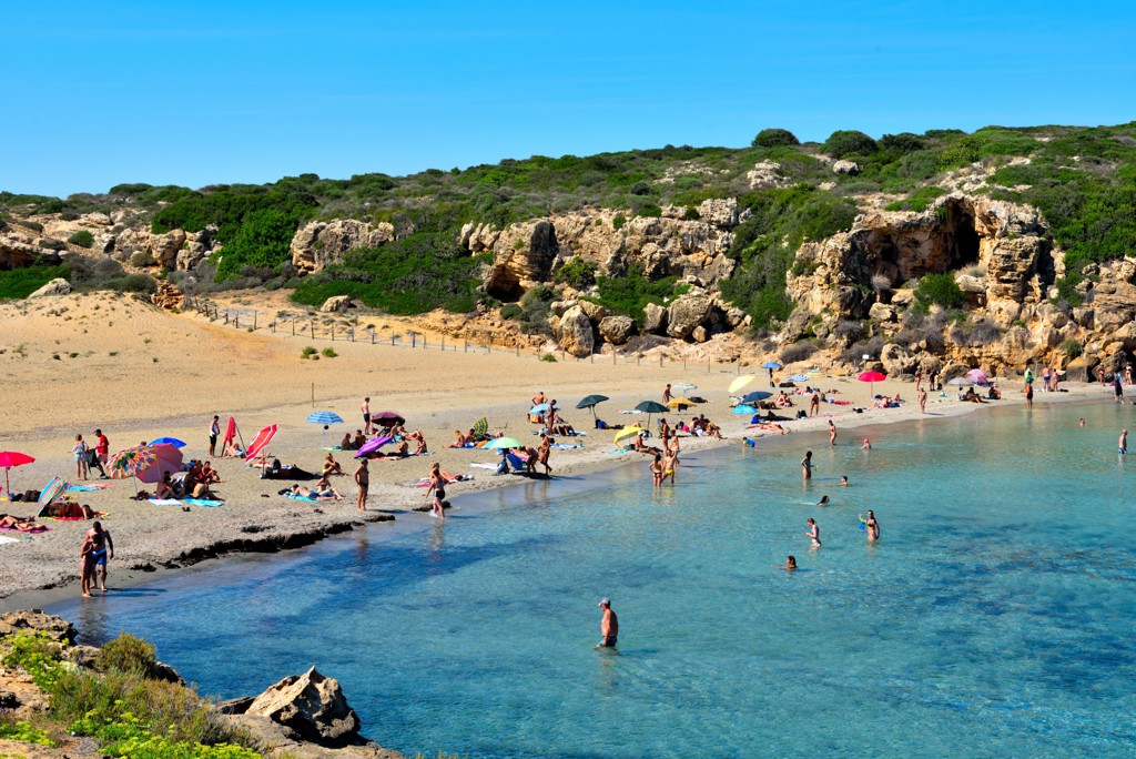 Zandstrand met helderblauw water en rotsachtige kust op Sicilië, met badgasten en parasols.