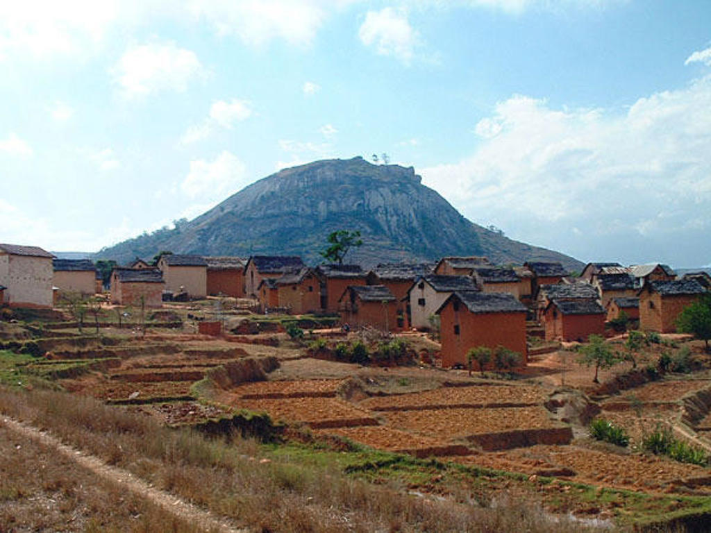 Traditioneel dorp met roodbruine huizen en terrassen in het landschap van Madagascar, met een rotsberg op de achtergrond.