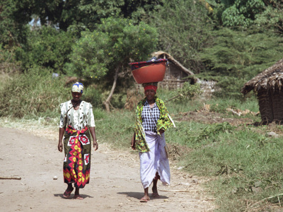 Twee vrouwen lopen over een zandpad langs hutten in een dorp in Tanzania.