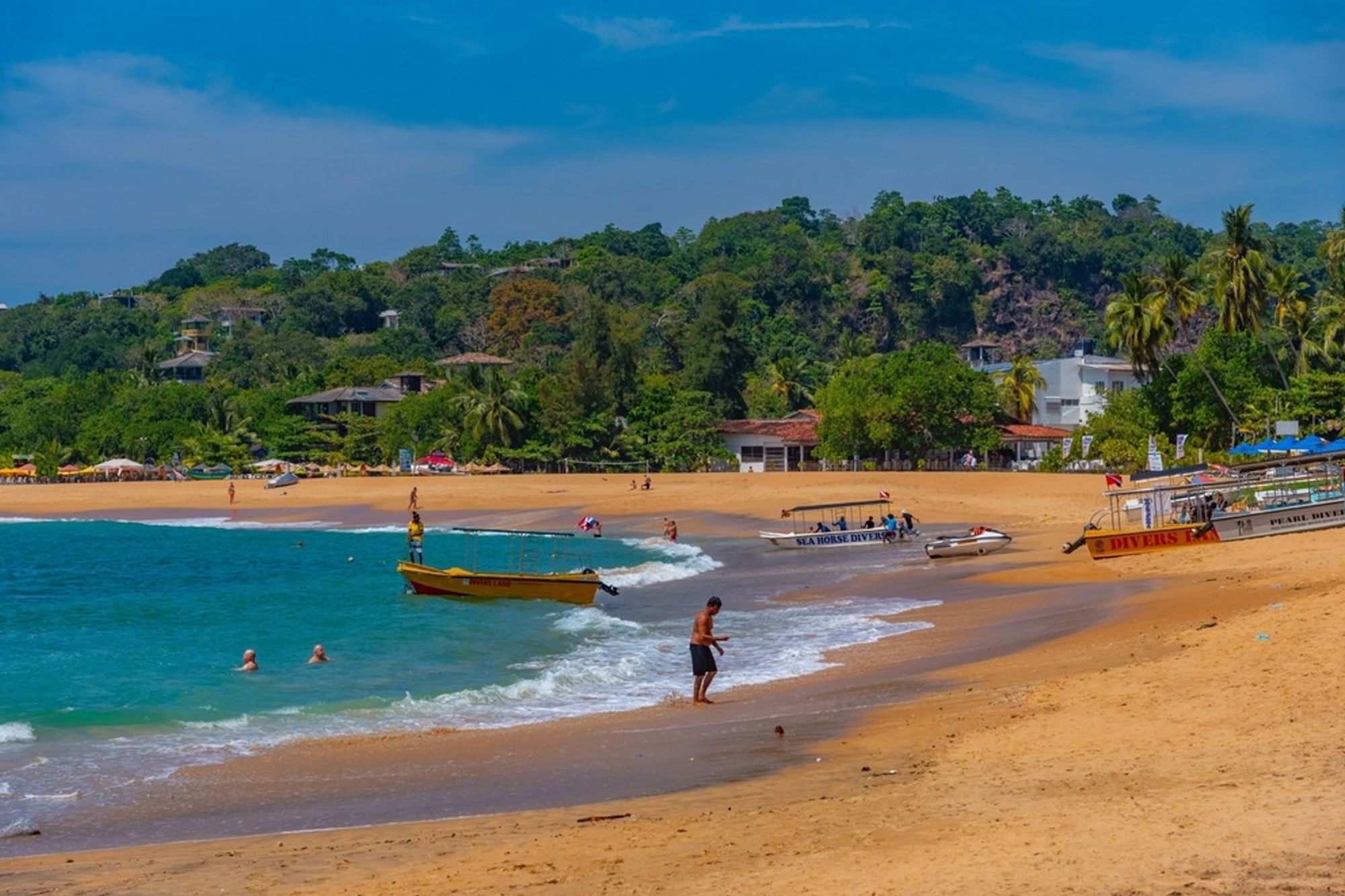Zandstrand in Sri Lanka met turquoise zee, vissersboten aan de kust en groene heuvels op de achtergrond.