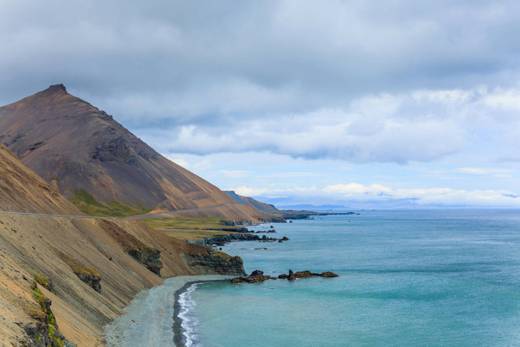 Kustlandschap in IJsland met steile berghelling, stille baai en turkoois zeewater onder bewolkte lucht.