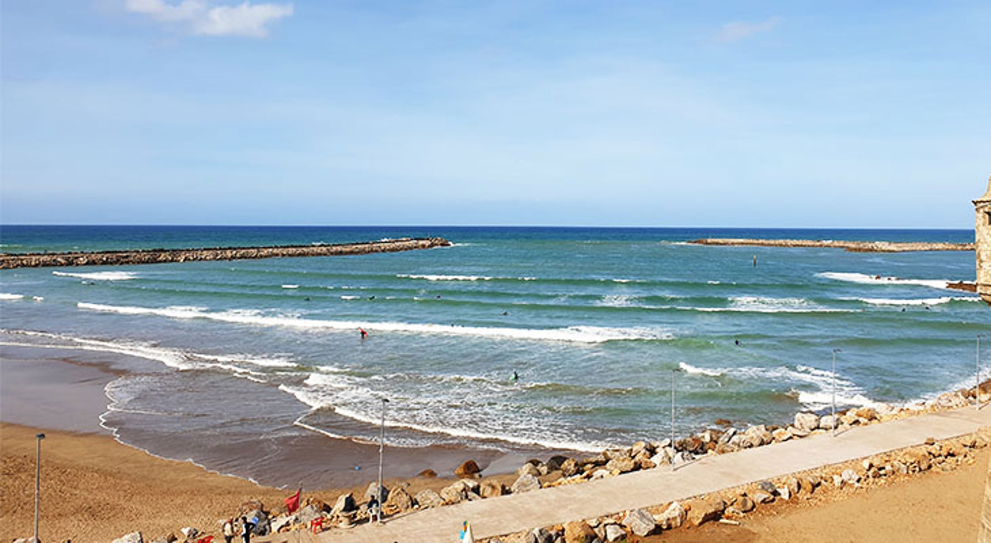 Uitzicht op strand en zee met golven, surfers en golfbrekers onder een heldere blauwe lucht.