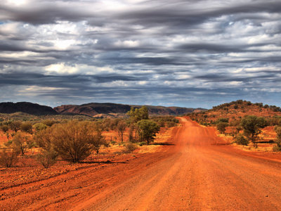 Rode onverharde weg door de Australische outback met lage heuvels en dreigende wolkenlucht.