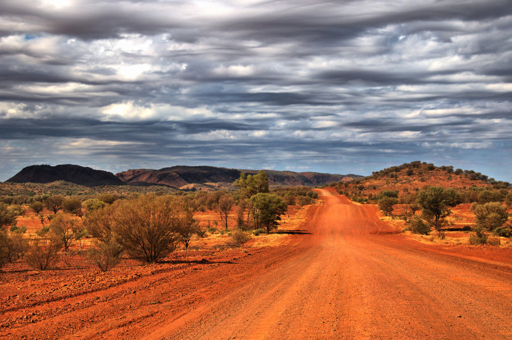 Rode onverharde weg door de Australische outback met lage heuvels en dreigende wolkenlucht.