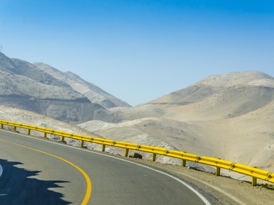 Bochtige bergweg in Peru met gele vangrail en uitzicht op kale bergen onder een heldere blauwe lucht.