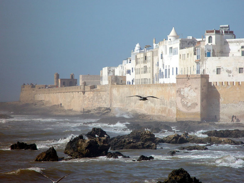 Stadswal en witte huizen aan de kust van Essaouira, Marokko, met rotsen en golven in zee.