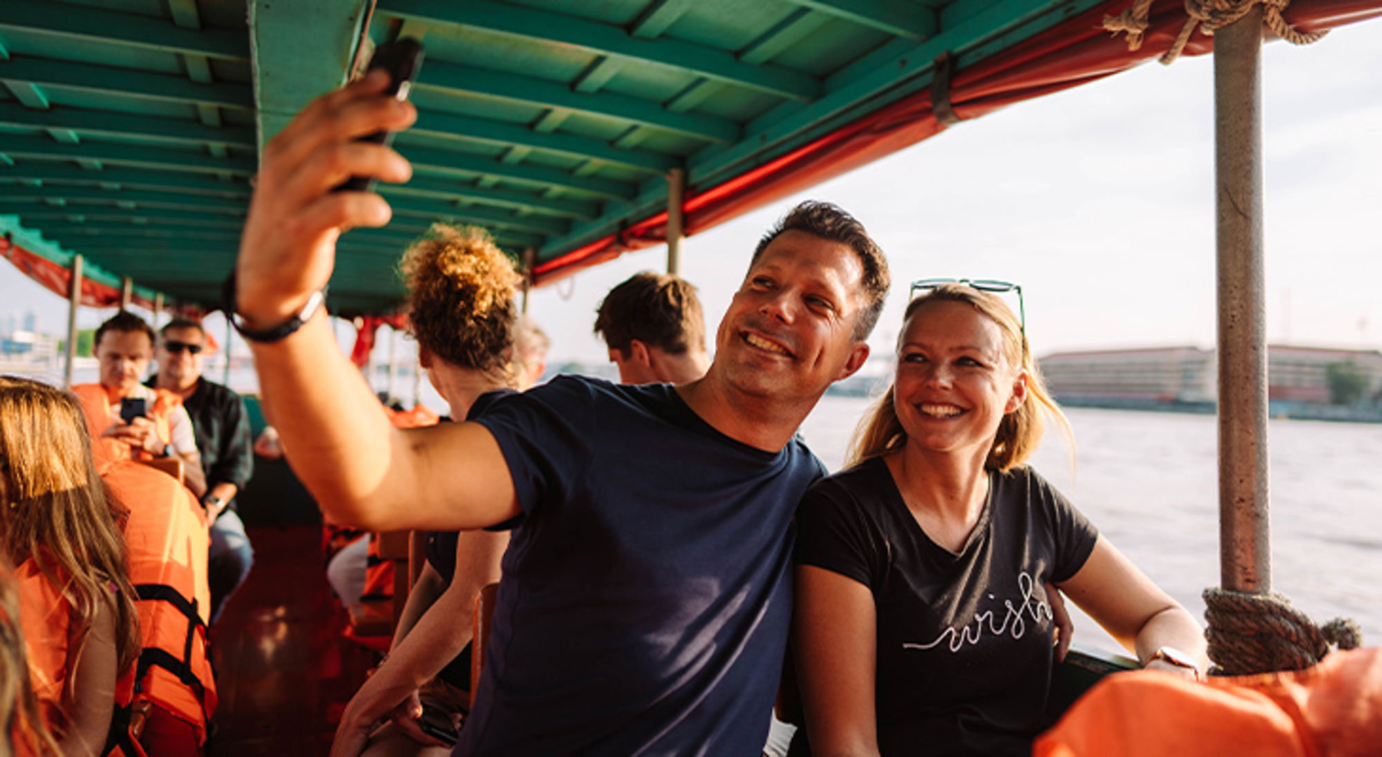 Stel neemt selfie op een boottocht, met passagiers in reddingsvesten op de achtergrond.