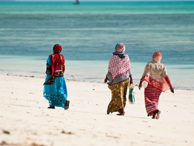 Drie vrouwen in kleurrijke doeken wandelen langs het witte strand van Zanzibar tijdens de route Kenia, Tanzania en Zanzibar.