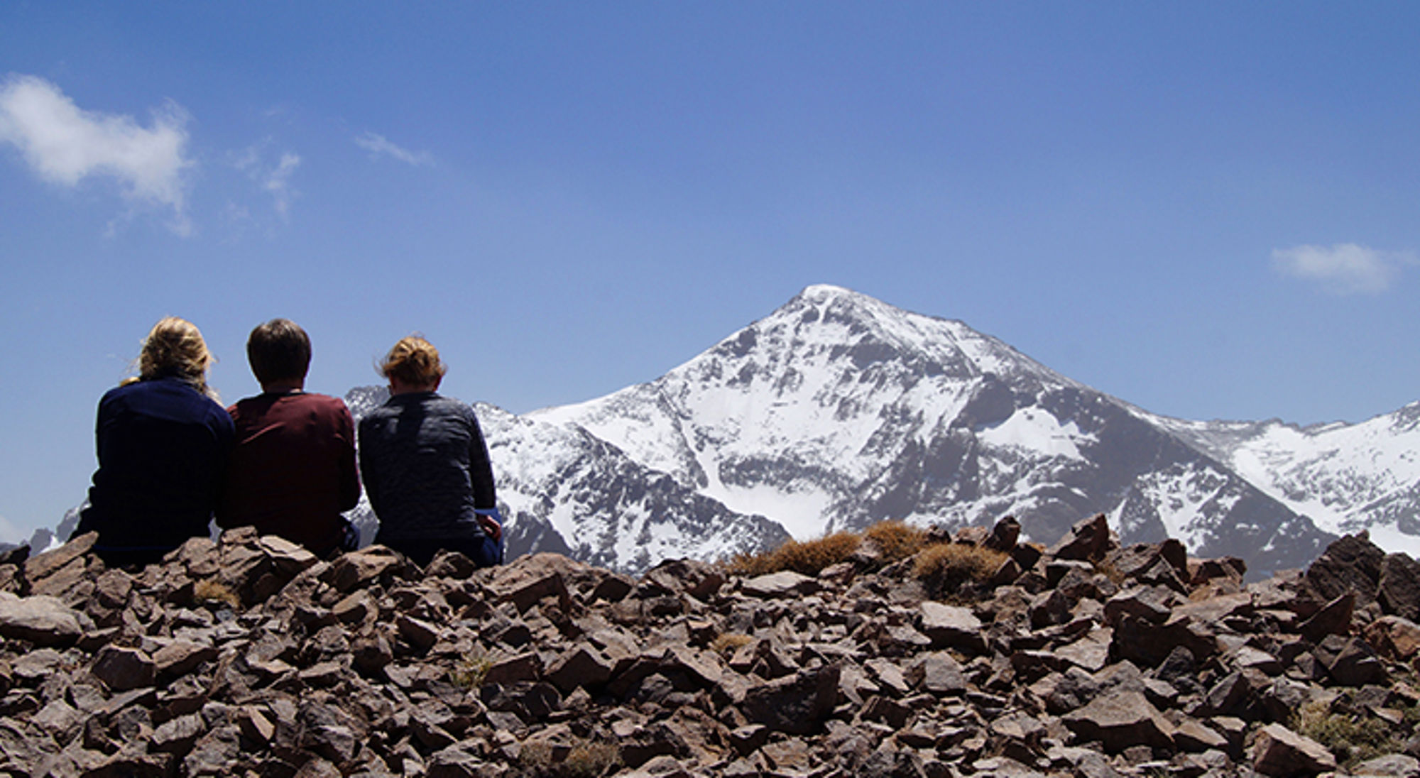 wandelvakantie marokko Drie wandelaars zitten op een rotskam en kijken uit op een besneeuwde bergtop onder een heldere blauwe lucht.
