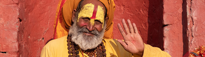 Sadhu met gele hoofddoek en gezichtsbeschildering groet voor een rode muur in Nepal.
