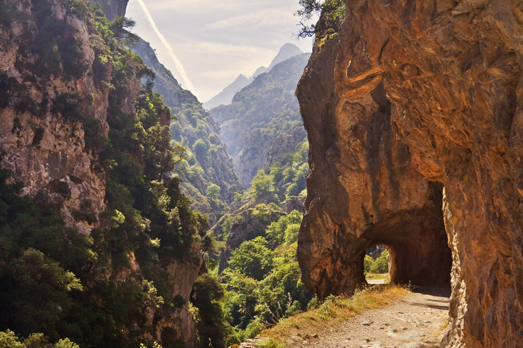 Wandelpad door een rotstunnel in de Picos de Europa, Spanje, met uitzicht op een groene kloof en bergen.