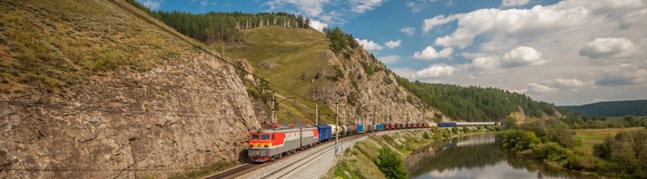Trans-Siberië Expres langs een rivier met rotswanden en groene heuvels onder een blauwe lucht.