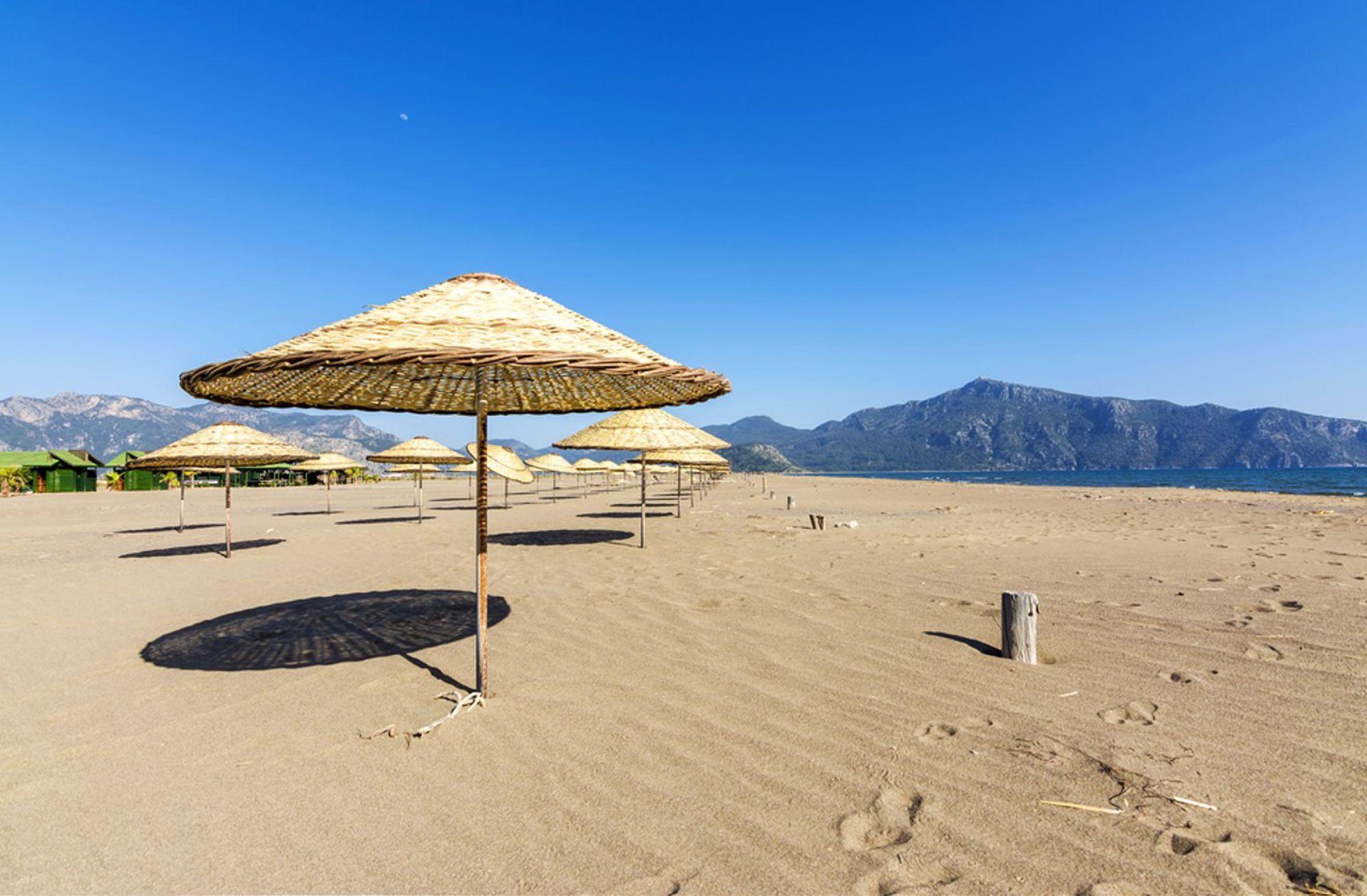 Rieten parasols op een rustig zandstrand aan de Turkse kust, met bergen op de achtergrond.