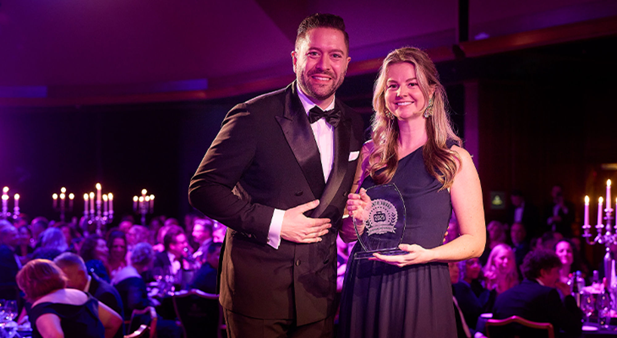 Man en vrouw in galakleding poseren met een award tijdens een prijsuitreiking in een feestzaal.