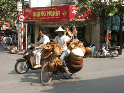 Straatbeeld in Hanoi: fietser met rieten manden en hoeden tussen het verkeer.