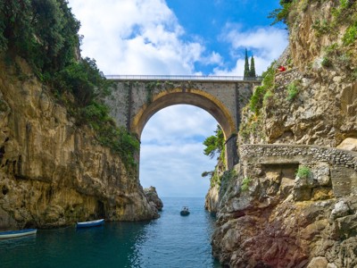 Boogbrug over smalle zeearm tussen rotskliffen aan de Amalfikust in Italië, met bootjes op helderblauw water.
