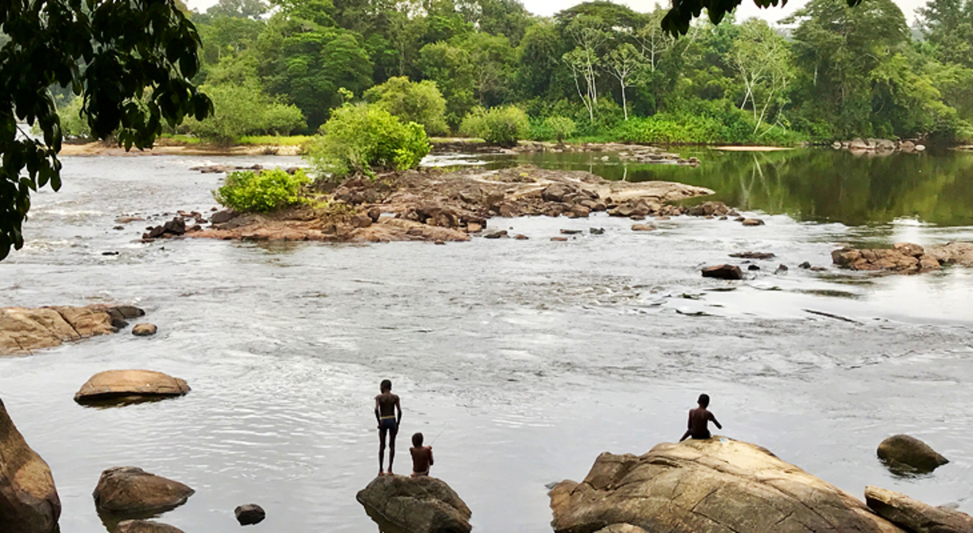 Kinderen spelen op rotsen in een brede rivier met groene jungle op de achtergrond.